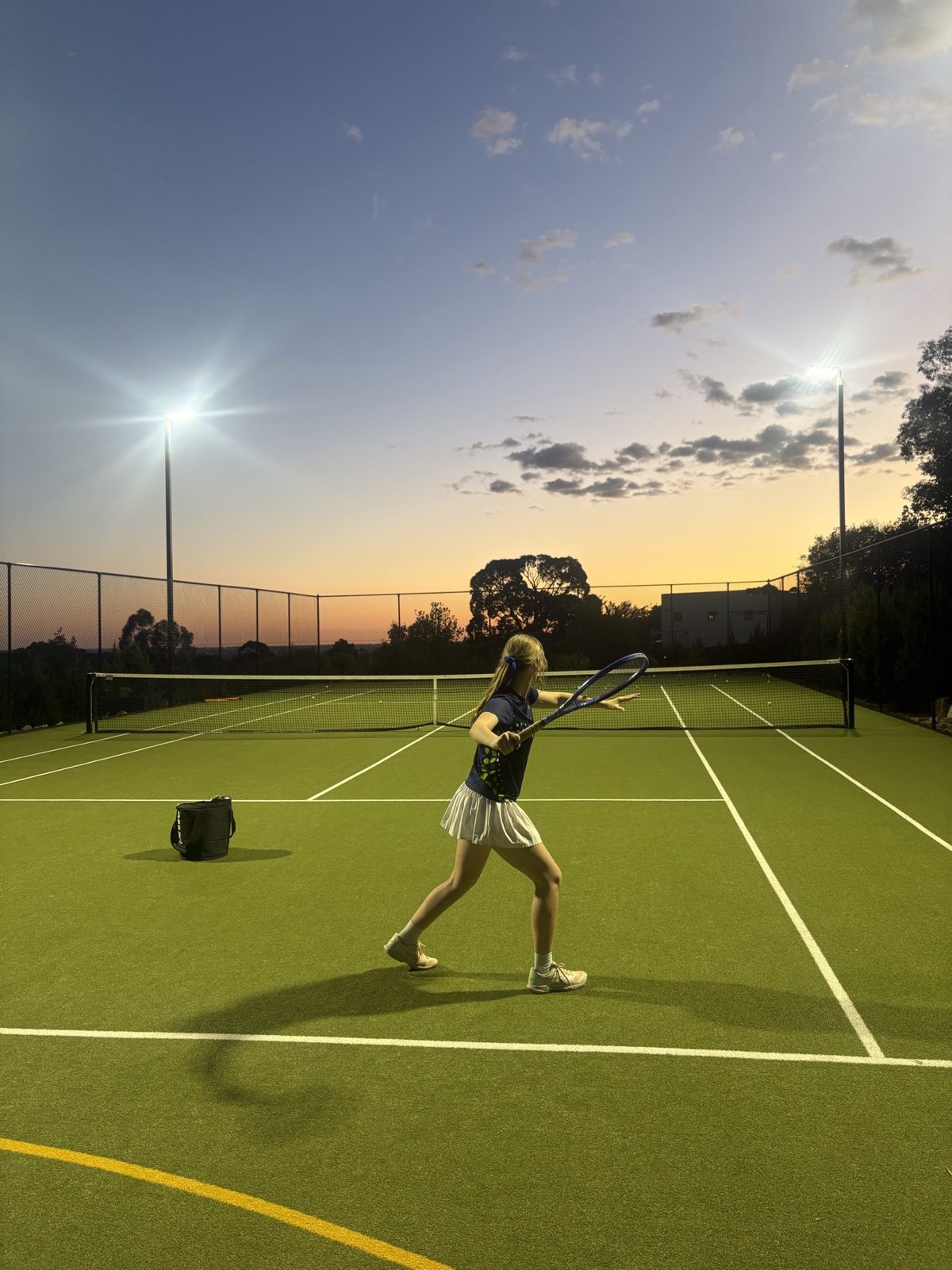 A junior player finishing a forehand on the floodlit court at dusk